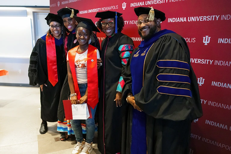 Group of five individuals dressed in Indiana University graduation gear pose in front of IU wall art