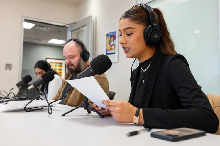 Three people sit at a table wearing headphone with microphones on small stands sitting on the table in front of them. The woman closes holds a pieces of paper and speaks into the microphone.