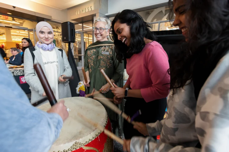 Students and staff gather around a drunk participating in a group drum beat at an international event in the Campus Center.