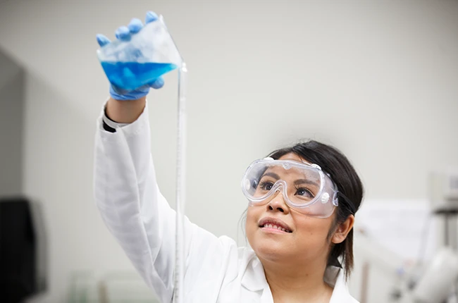 A student in a lab coat and safety goggles pouring a blue liquid from a beaker
