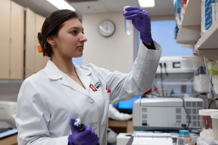 A woman wearing a School of Medicine lab coat holds up a test tube to look at the solution inside. Lab equipment can be seen in the room behind her.
