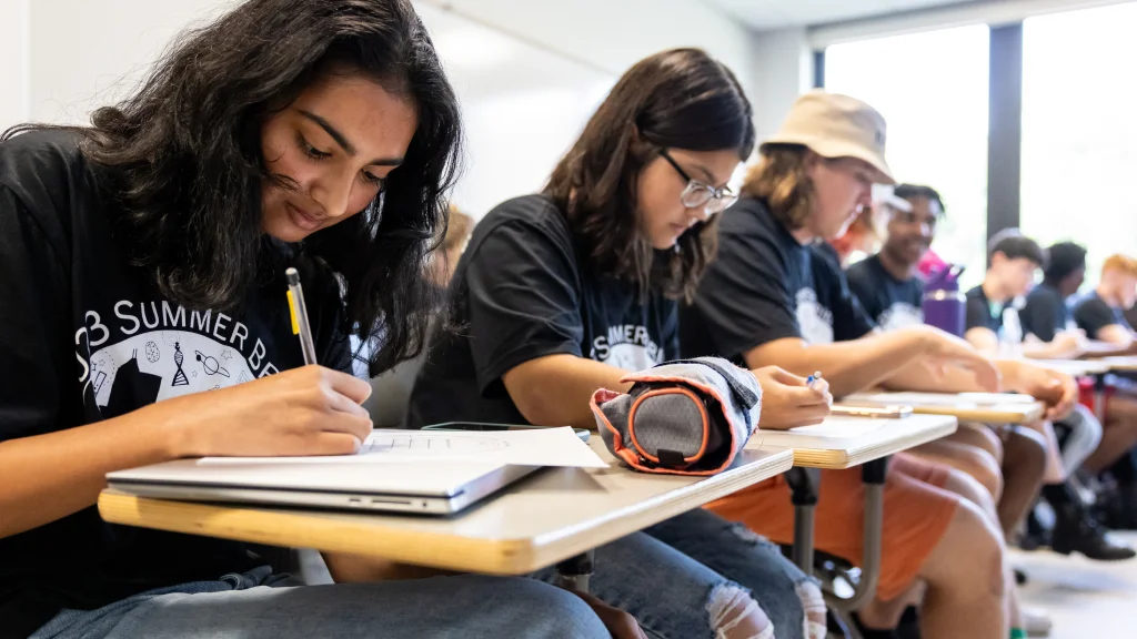A row of several students sit at desks in a bright classroom looking down and writing on a paper in front of them.