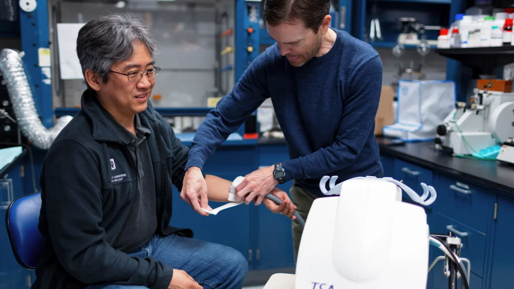 A young man attaches wiring to a sitting man’s hand, hooking him to nearby machinery that will collect data.