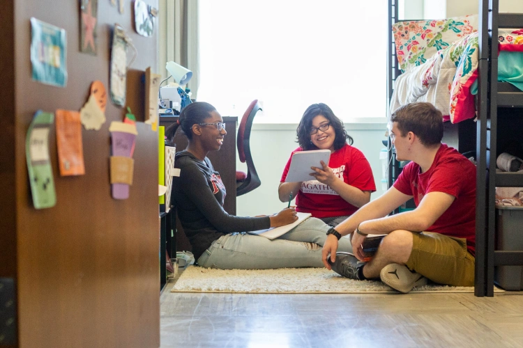 View through an open residence hall door to a room where three students are sitting on the floor working on a project, holding notebooks in their lap.