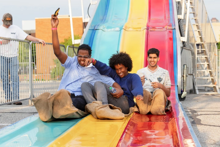 Three students sitting on burlap sacks slide to the bottom of a carnival slide set up for the end of year Jagapalooza celebration.