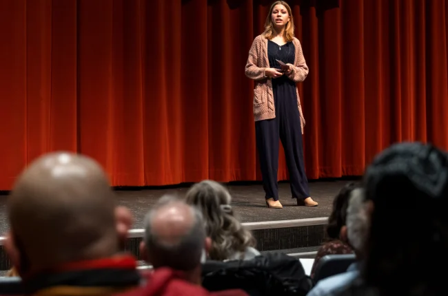 A woman stands on a stage with a red curtain speaking to a seated audience.