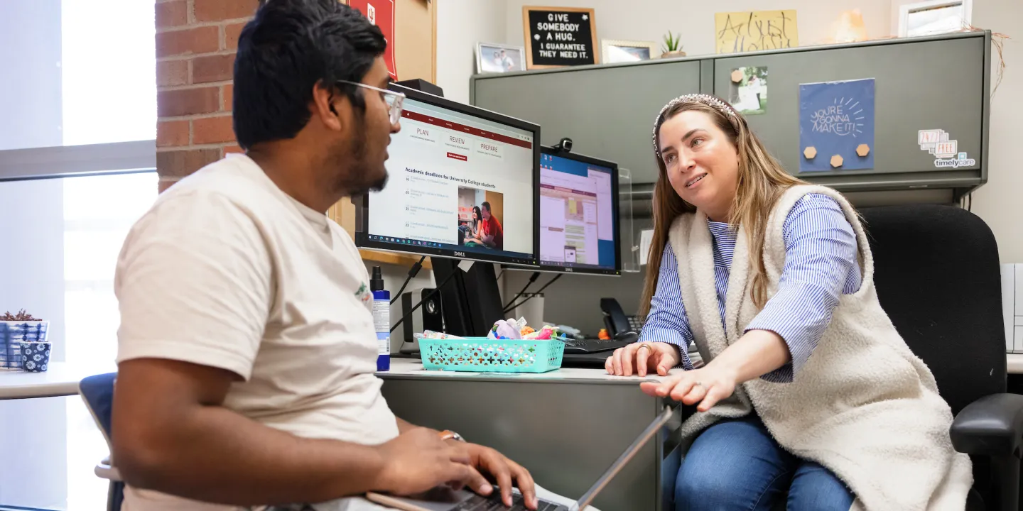 A student sits at an advisor’s desk with a laptop on his lab. She is motioning to his laptop and speaking to him.