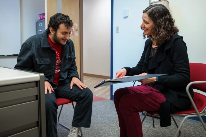 A student sits with an advisor on red chairs in and office. The advisor holds out and points to paper inside a binder. The student had his hands on his knees and is smiling. 
